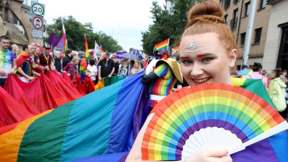 A marcher holds a rainbow fan next to a rainbow flag running down the street behind them.