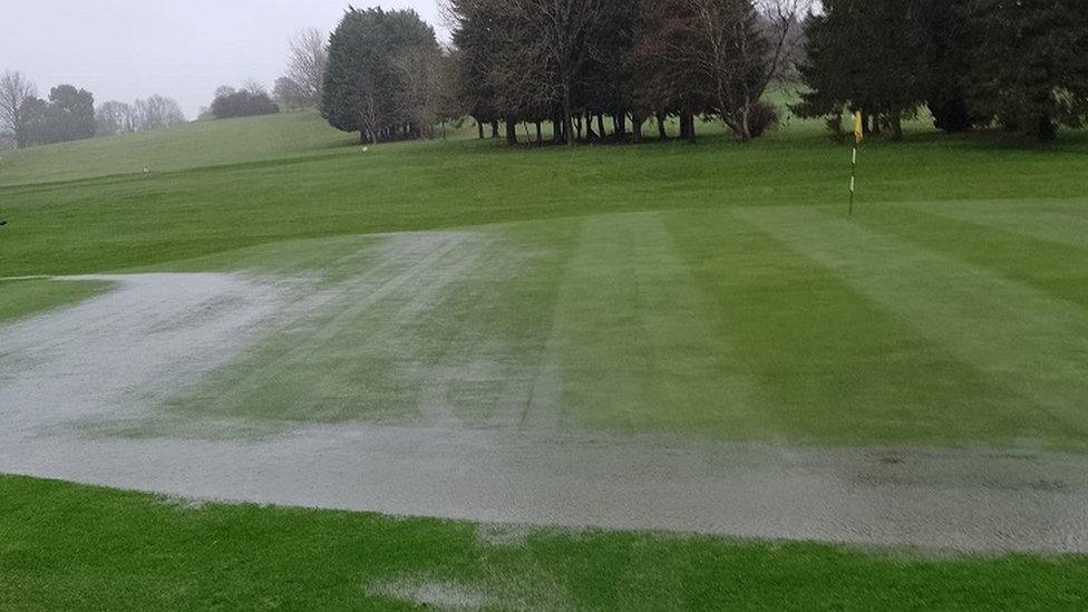 Water across the grass at a golf club with trees in the background.