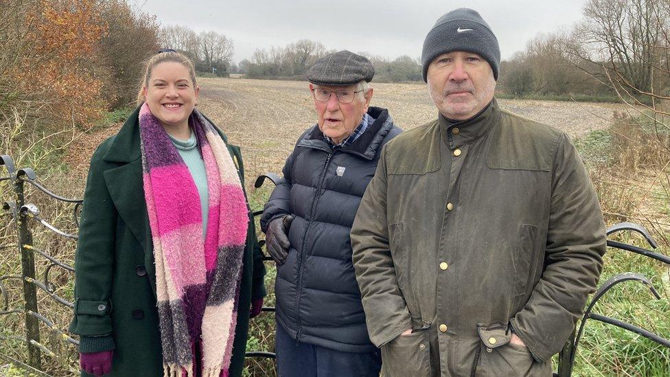 Three people standing in front of the land in winter coats - one wearing a thick colourful scarf. The land is showing it is winter, with leaves off the trees and the field bare.