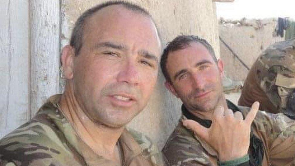 Two men in military uniforms pose for the camera, leaning against a wall in an arid location.