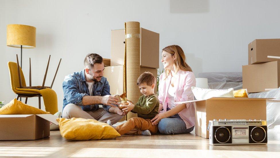 A couple and their young son sit in a living room surrounded by moving boxes and furniture.