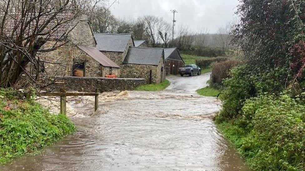 Water floods a country road with green verges to each side and low buildings and a car at the bottom