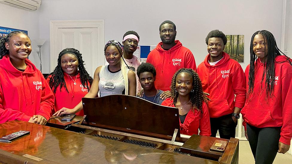 Nine members of the Reggae Roots choir are gathered, smiling into the camera, around a piano in a rehearsal room. Most of them wear red hoodies with the group's name printed on it.