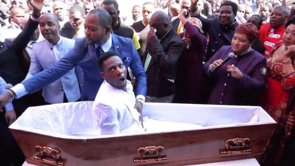 Pastor Alph Lukau stands next to the coffin and places his hand on the stomach of the man he is claiming to resurrect.