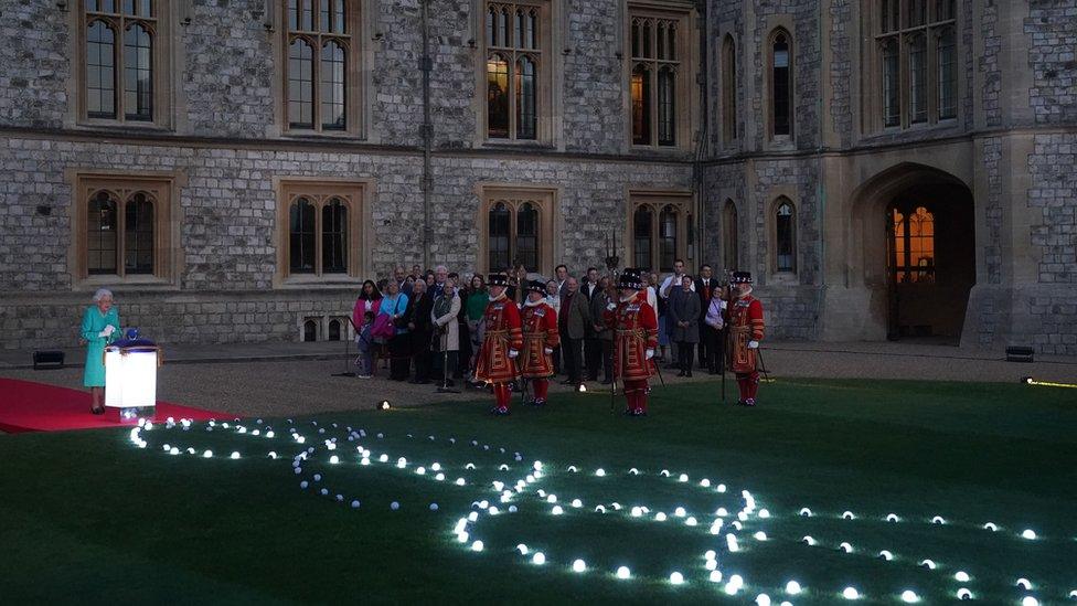 the queen stands in a courtyard of windsor palace with guards standing by as she lights a beacon