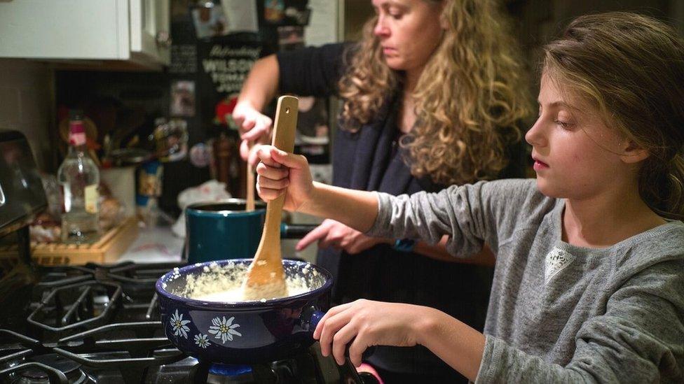 A child stirs a pot on a hob as he mum cooks beside her