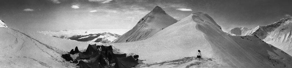 A wide-angled shot shows small tents nestled in a group between snow-covered peaks