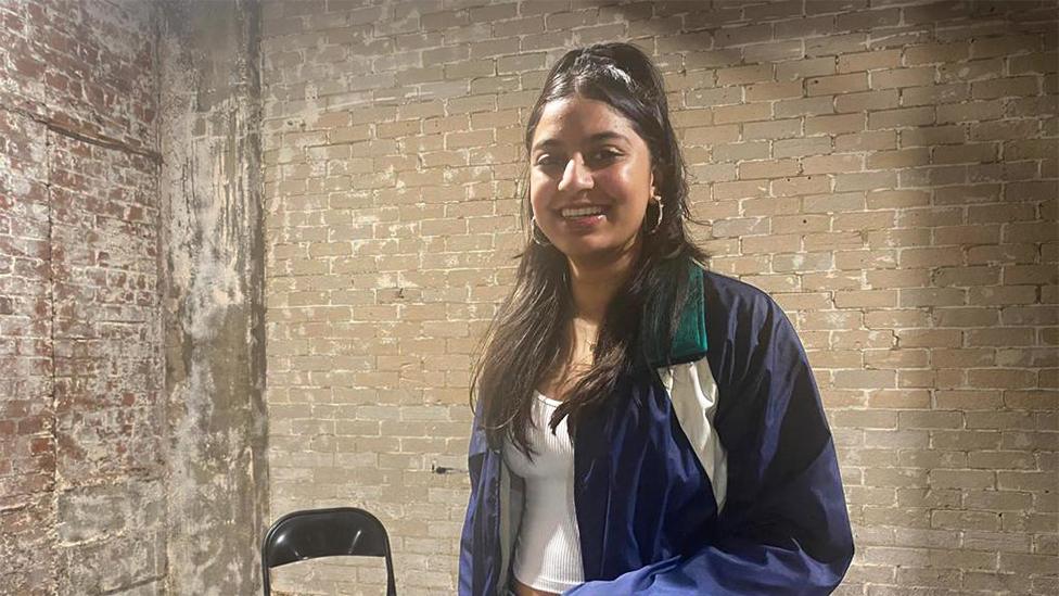 Siyani smiling at the camera, wearing hoop earings, a navy blue jacket and a white top. The background is a discoloured brick wall, with white marks. There is a foldable black chair behind Siyani