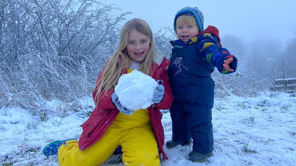 A girl holds a big snowball and poses with baby brother