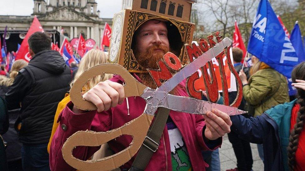 A man holds a cardboard placard that reads: NO MORE CUTS