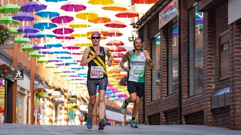 A man and woman run beneath a canopy of rainbow coloured umbrellas