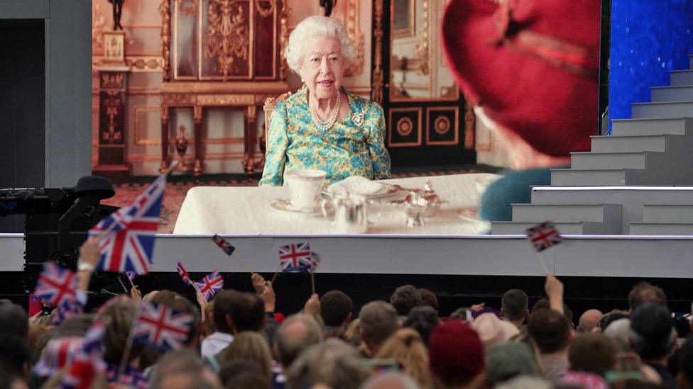 The Queen and Paddington Bear on a big screen, with people waving union flags in the foreground