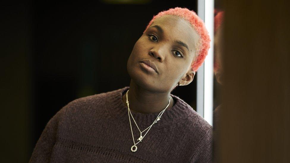 Arlo Parks looks straight into the camera with her head tilted slightly to the right. She has cropped pink hair. She's wearing a black fluffy jumper and some silver necklaces.