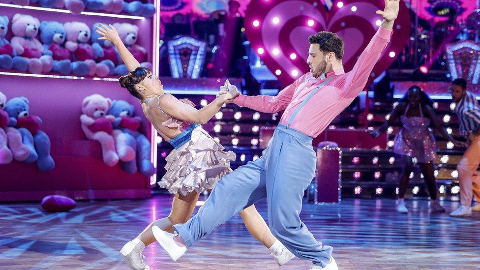 Ellie Leach and Vito Coppola during one of their performances. The set is bubblegum pink, staged with multiple pink and blue teddy bears and pink hearts. Ellie wears her brown hair tied back in a bun with a pink and blue ruffled dress while Vito is wearing blue trousers and a pink shirt. In their routine, they hold hands while leaning back, their free arms outstretched behind them