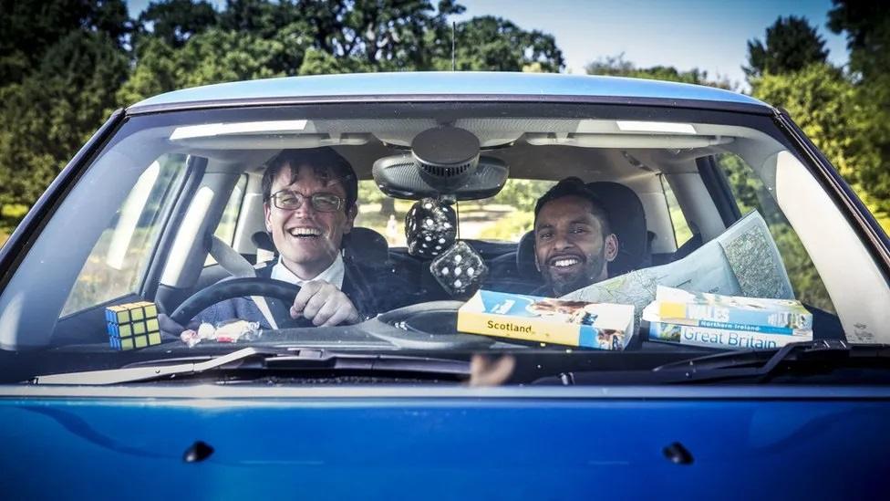 Man with short hair and glasses and man with short hair and beard laughing in a car