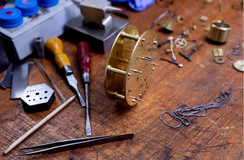 A close-up of clock parts and hand tools on a work bench