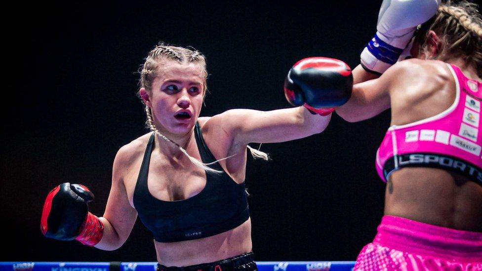 Daniella Hemsley, a blonde white woman with her long hair in braids wears a black sports bra as she aims a punch at opponent Aleksandra Daniel at their fight in Dublin on Saturday 15 July. Aleksandra Daniel wears a pink sports bra with matching shorts and stands with her back to the camera, covering her face with blue and white boxing gloves to defend against Daniella's jab