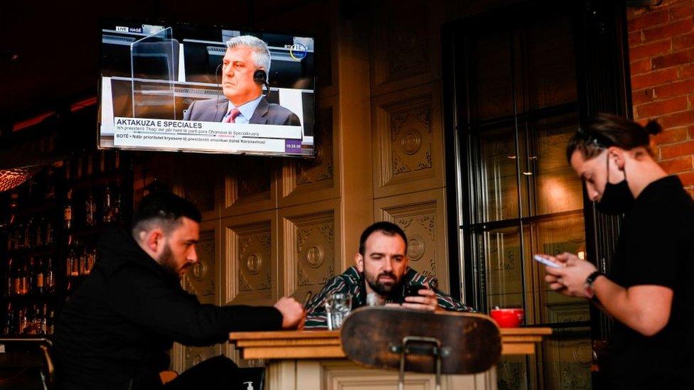 Former president Hashim Thaci is seen on a TV as people look at their mobile phones in a coffee bar in Pristina. Thaci is appearing before a Hague war crimes court in The Hague on November 9, 2020, to face charges relating to the 1990s conflict with Serbia.