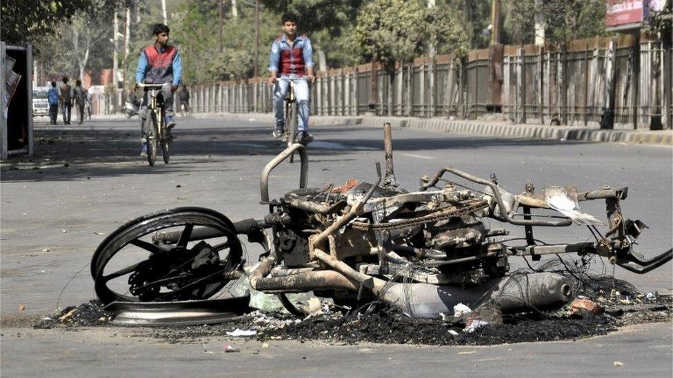 People ride their bicycles close to a damaged motorcycle set alight by protesters during a demonstration by members of Jat community in Rohtak, in the northern state of Haryana, India, February 21, 2016