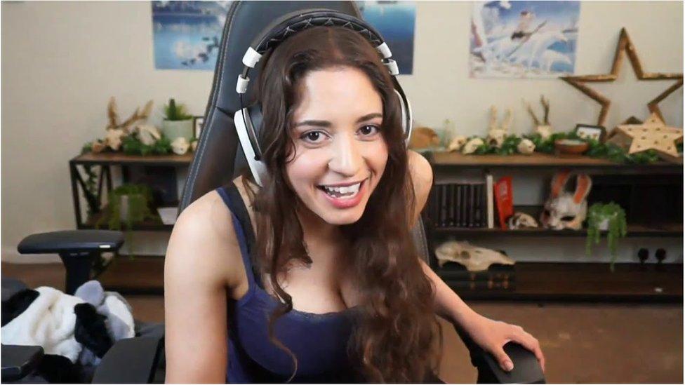 A photo of anita sat in a gaming chair with a headset on, she is smiling and laughing whilst looking into the camera with a bookshelf and posters in the background.