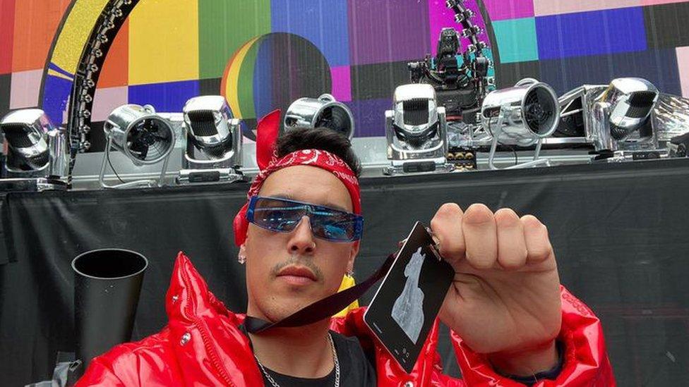 Bilal Rouabah in front of the stage at Beyonce's Renaissance tour. He's wearing a red coat and red bandana and blue-tinted sunglasses and his holding up a lanyard that's around his neck with the tour logo on it to the camera