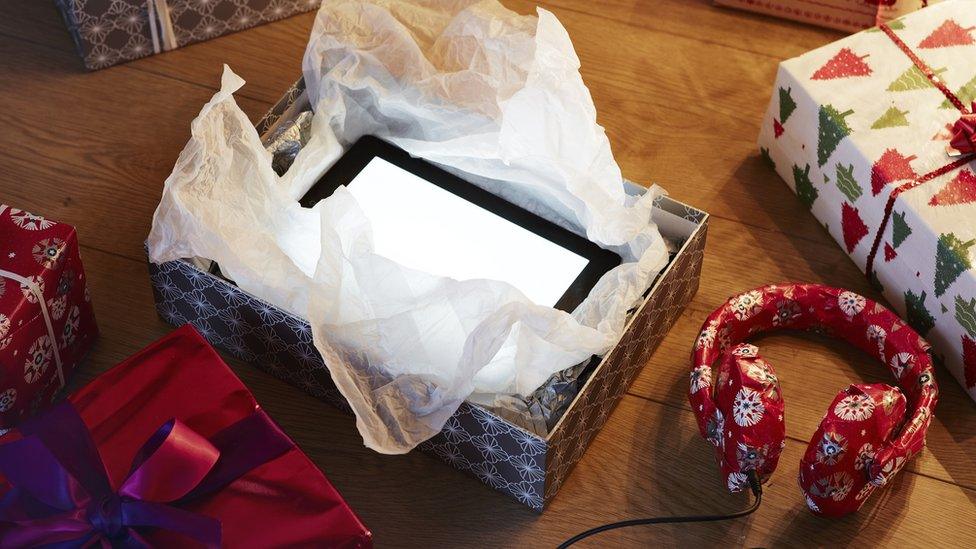 A tablet is being opened from a Christmas gift box, with more gifts seen nearby