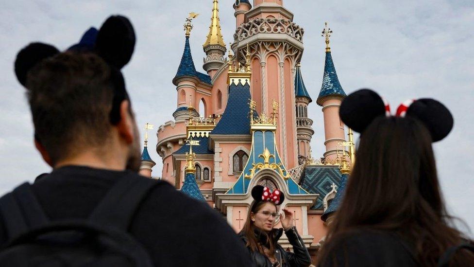 Visitors wearing emblematic Mickey and Minnie Mouse ears look on, in front of the Sleeping Beauty-inspired castle at Disneyland Paris, in Marne-la-Vallee, east of Paris, on 16 October