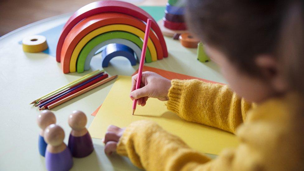 File image showing a young child drawing on paper next to wooden people and a stacking rainbow arch