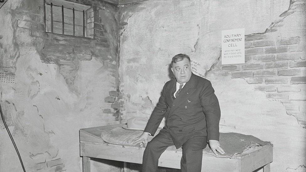 Black and white photo showing a middle-aged man dressed in suit and tie sitting on a basic bed in a room with brick and stripped off plaster and a small window with bars