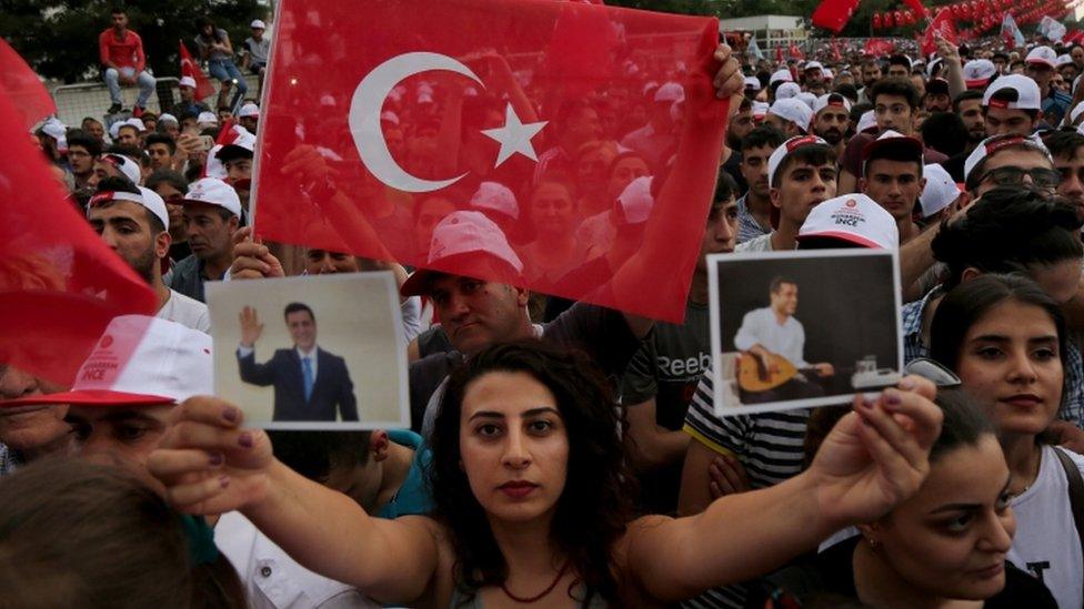 A woman at a rally surrounded by red Turkish flags holds up photographs of Selahattin Demirtas