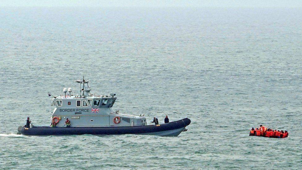 A grey Border Force boat faces a small orange dinghy full of people in the sea.