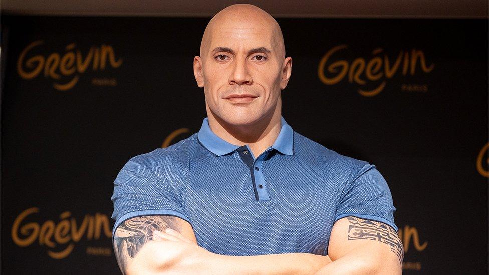 The Dwayne Johnson wax figure at Musee Grevin in France. The figure shows a likeness of Johnson in a blue polo shirt and tattoos on his arms.