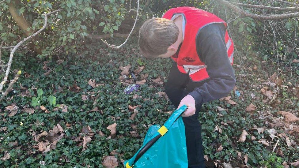 Boy with hi-viz vest carries a green back and is picking up a crisp packet