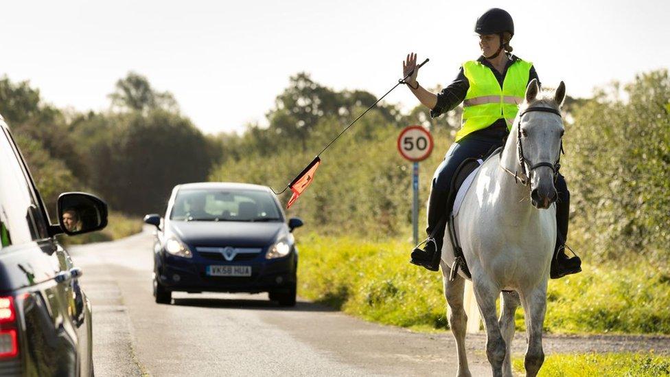 A woman in a high vis jacket riding a horse holds out the VisiWhip to an oncoming car to thank them for allowing a safe distance between them and her horse