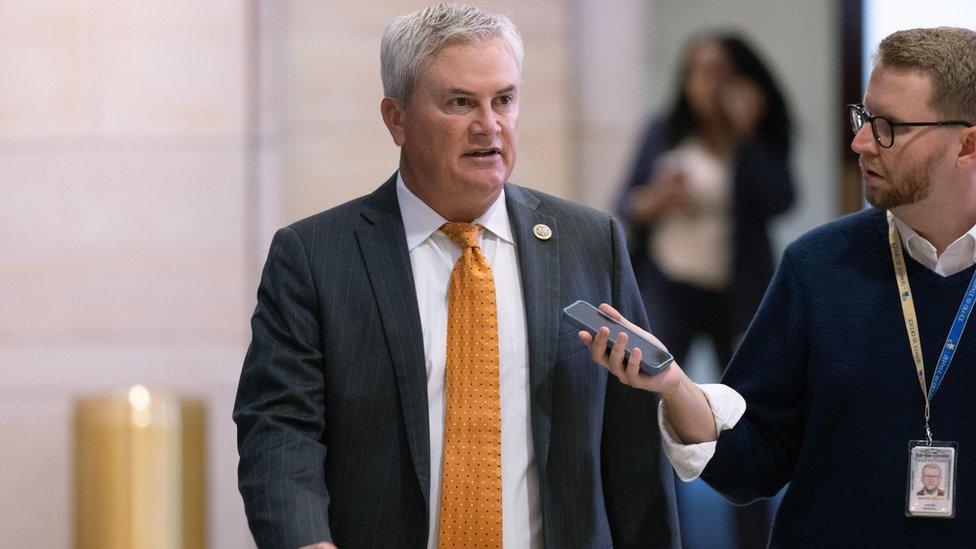 James Comer in yellow tie talking to reporter in hallway.