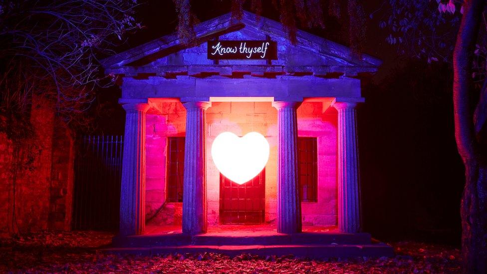 A bright white heart shines and appears to float in midair in a building that has purple and pink light illuminating it. On the top of the building are the words 'Know Thyself'.