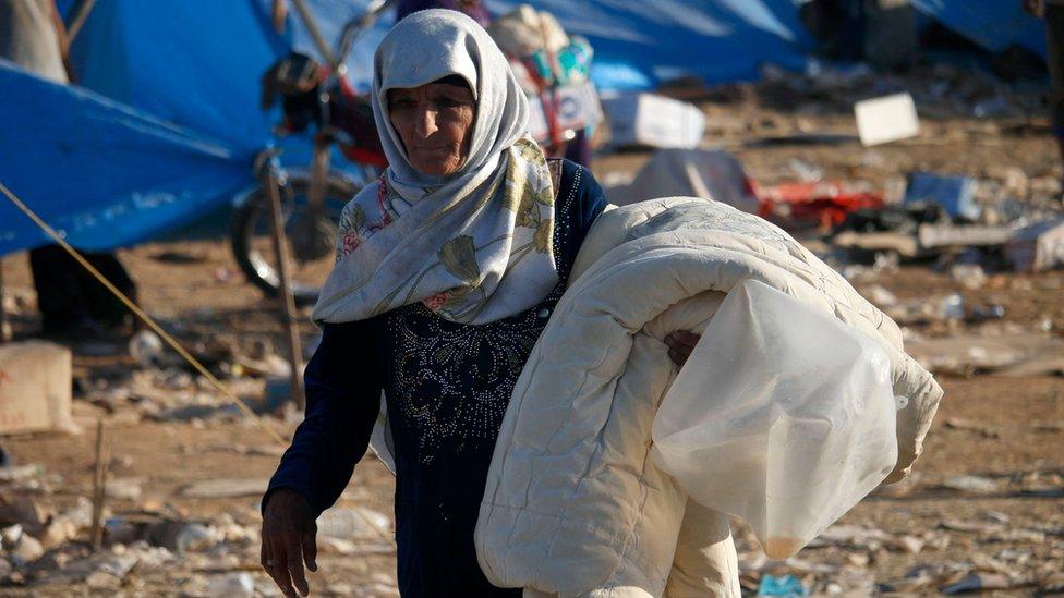 An elderly Syrian woman carries a duvet at a makeshift camp near the Nassib border crossing with Jordan (2 July 2018)