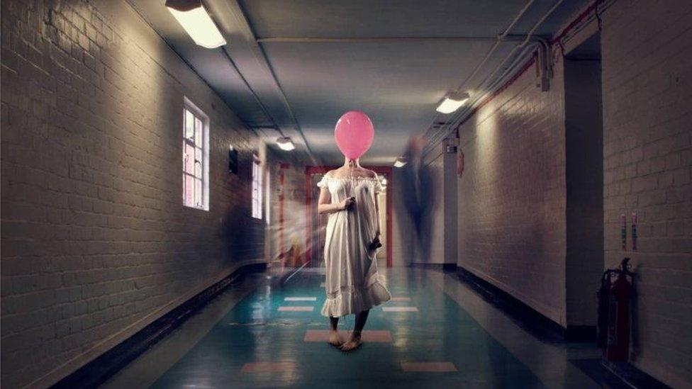 A woman in a corridor wearing a dress and covering her face with a pink balloon