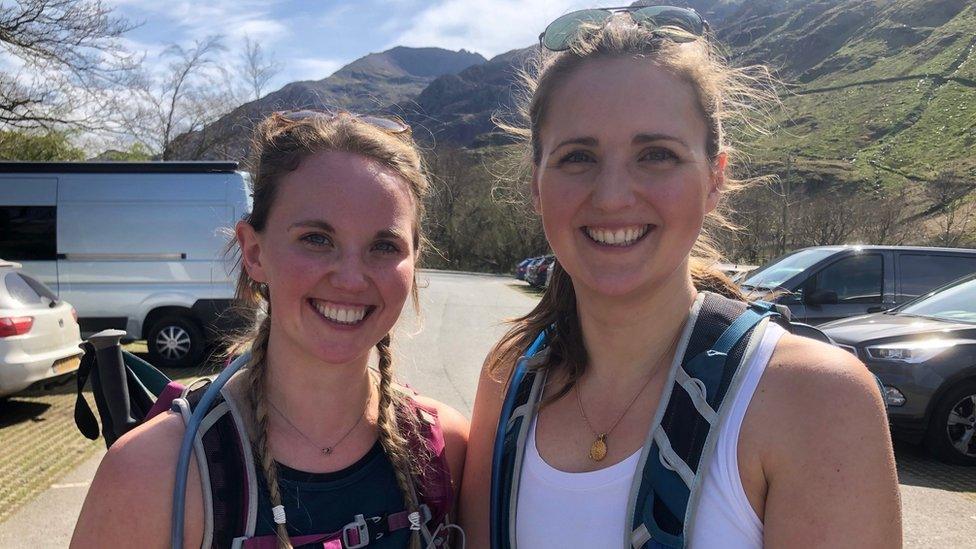 two women standing in a car park dressed in hiking gear with mountains in the background