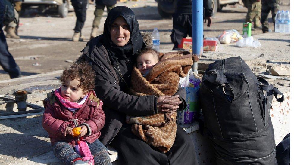 A mother cradles her baby while sitting next to her toddler and a suitcase