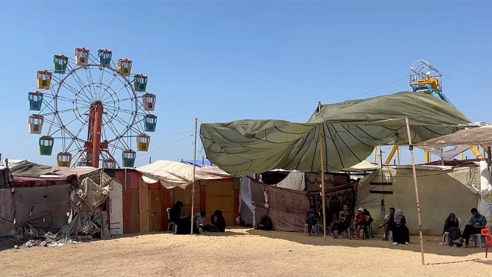 The Ferris Wheel seen in the last image can be seen again but now there are makeshift tents in the foreground with small clusters of people dressed in dark clothing sheltering beneath.