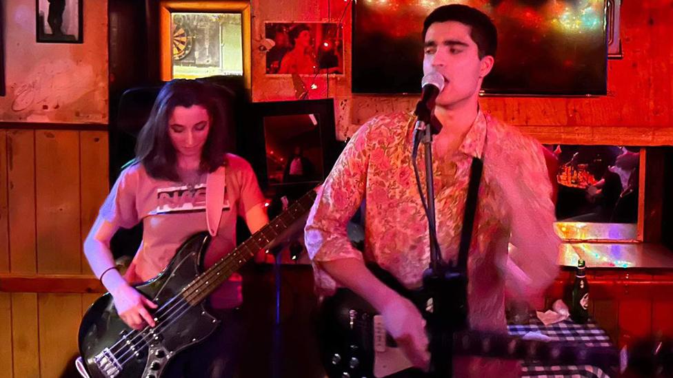 A man in a shirt with a pink floral pattern plays guitar and sings into a microphone on a small stage inside a bar or pub. Behind him a young woman in a nasa t-shirt with shoulder-length black hair plays a bass held over her shoulder by a thick pink strap. They're bathed in an orange-red stage light. A TV, mirrors of different sizes and two black and white pictures hang on the wall behind them.