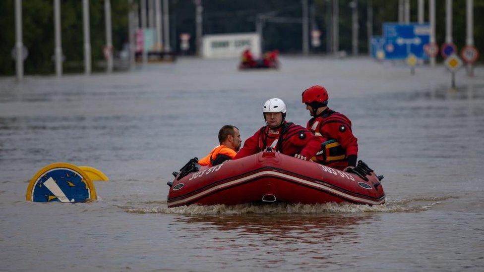 Two rescuers evacuate a flood-affected resident in a boat. The street is badly flooded, with road signs partly under water.