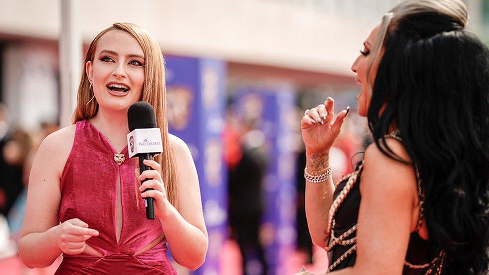 Amelia Dimoldenberg at the Bafta awards with Michelle Visage. Amelia is wearing a pink dress, looking to the right while holding a microphone in her left hand, with her right hand pointing towards Michelle. She is interviewing Michelle who is wearing a black dress with chains around the outfit, with her right hand gesturing. The background is blurred