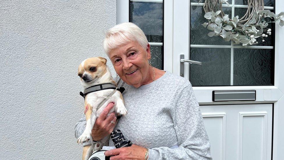 Park home resident Carol standing in front of her front door holding her small pet dog Barney