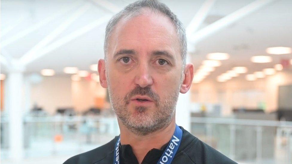 Man with short hair and beard, wearing a lanyard stands in a large building