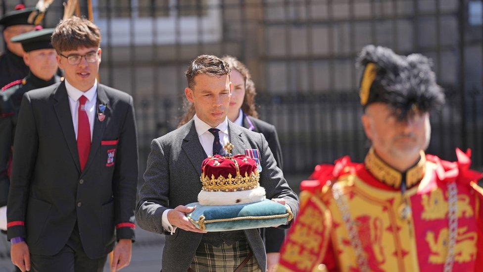 The Duke of Hamilton wears a suit and carries a blue pillow with the Crown of Scotland, followed by two pupils in school uniforms