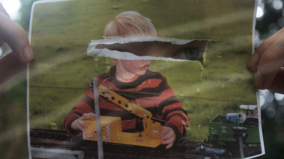 A boy is holding a photograph from his childhood in front of his face. He is blonde and playing with a toy truck. The photograph has been torn in the middle over his face. Through the hole, you can see the boy's blue eye peering through.