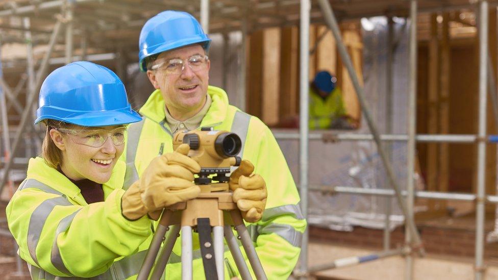 A female construction worker stands behind a builder's level on a building site