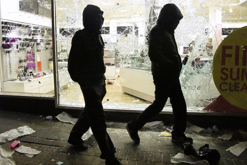 Two men with their faces covered walk past a looted shop during the riots in 2011